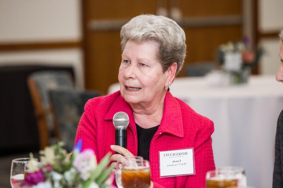 A woman holding a microphone while speaking at a dining table. She is wearing a bright pink jacket and a name tag for Agnes Scott College. She has short, gray hair and is seated in a room with other attendees in the background.